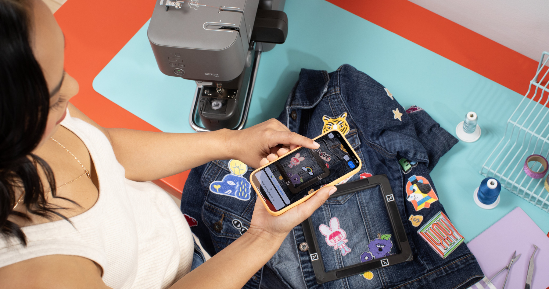 A person using Brother Artspira app to customize a denim jacket with patches and an embroidery machine in view.